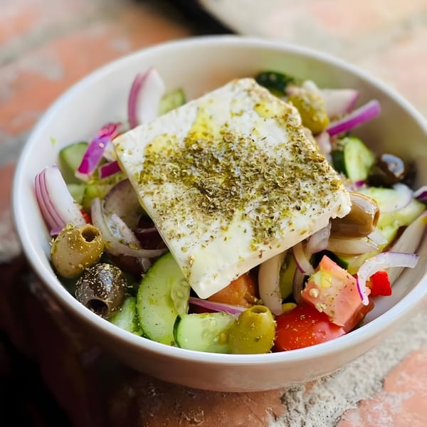 A Mediterranean salad with a large block of feta cheese, olives, cucumbers, tomatoes, and red onions.