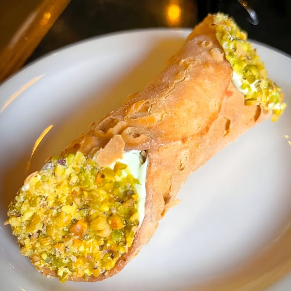 A single cannoli from Alpha Omega Grill & Bakery, filled with cream and topped with chopped pistachios.