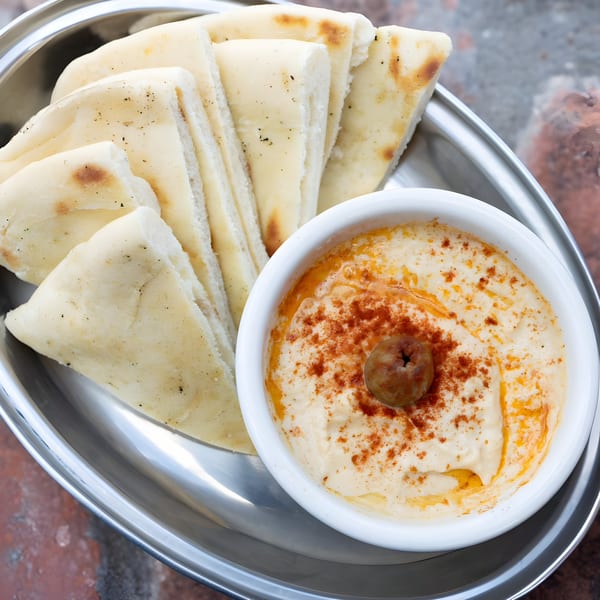 A metal platter holds a white bowl of hummus with paprika and an olive, alongside pita bread.